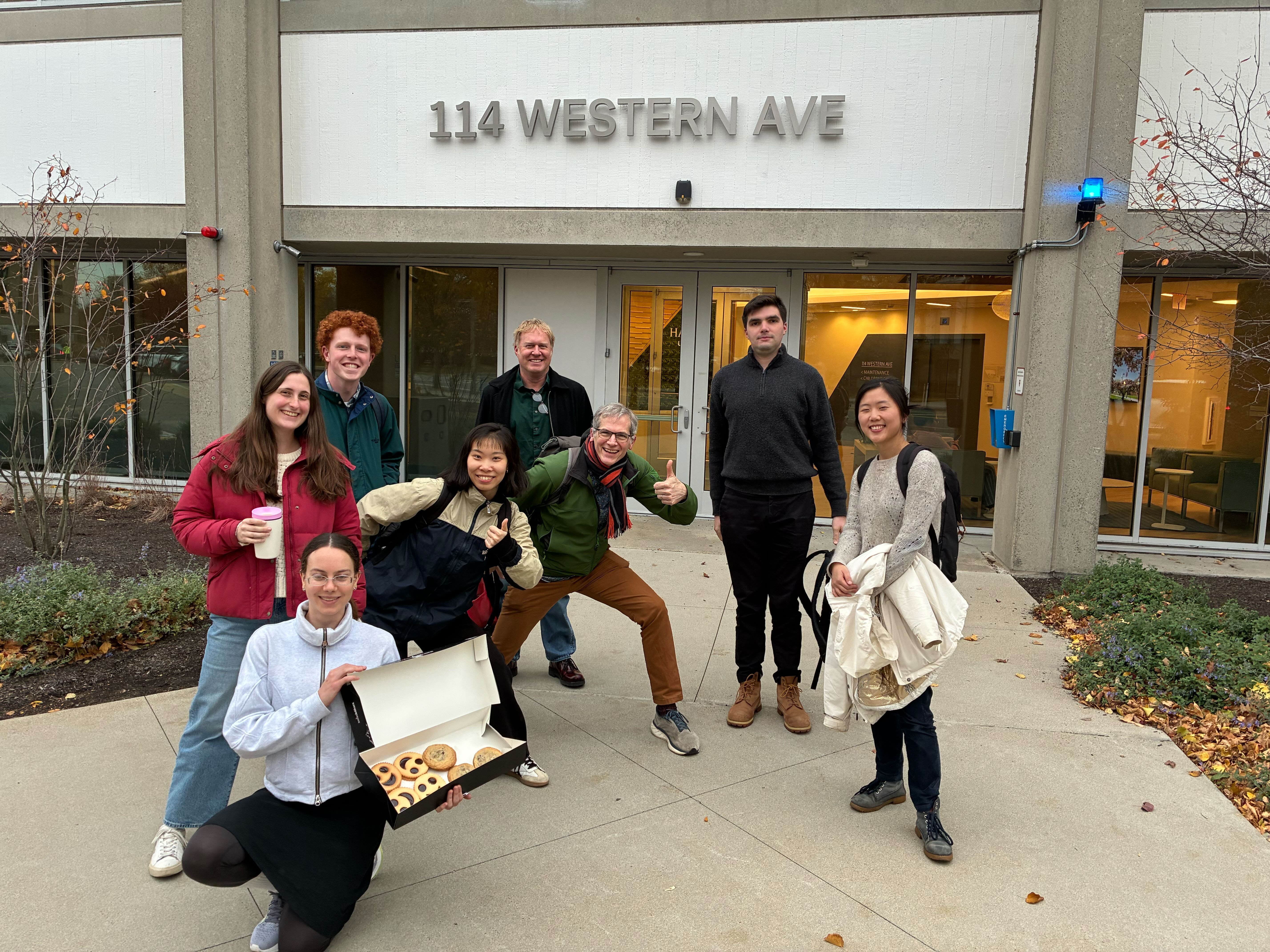 A group of people posing in front of an academic building, with one person showing off a box of cookies